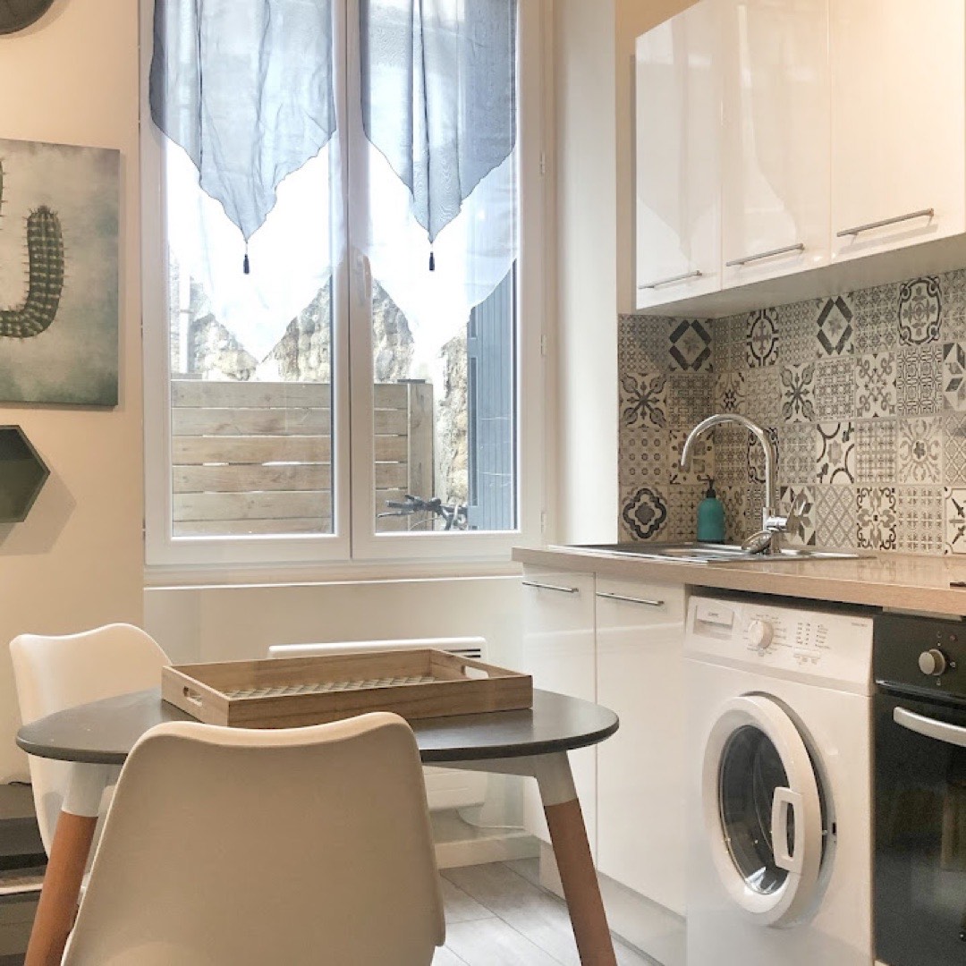 Cuisine équipée blanche d'un studio : crédence carreaux de ciment, plaque, four, lave-linge et coin repas près de la fenêtre.