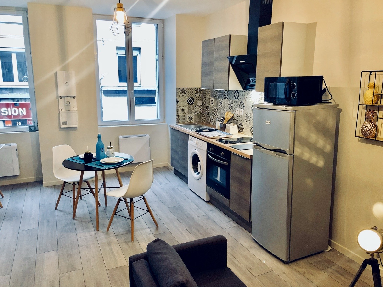 Pièce de vie lumineuse d'un studio meublé : coin repas, cuisine équipée et canapé près d'une grande fenêtre.